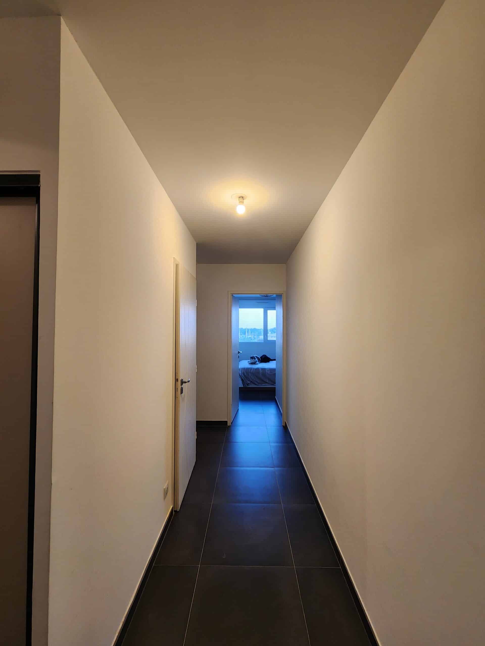Couloir moderne aux murs blancs et carrelage sombre menant vers une chambre lumineuse.