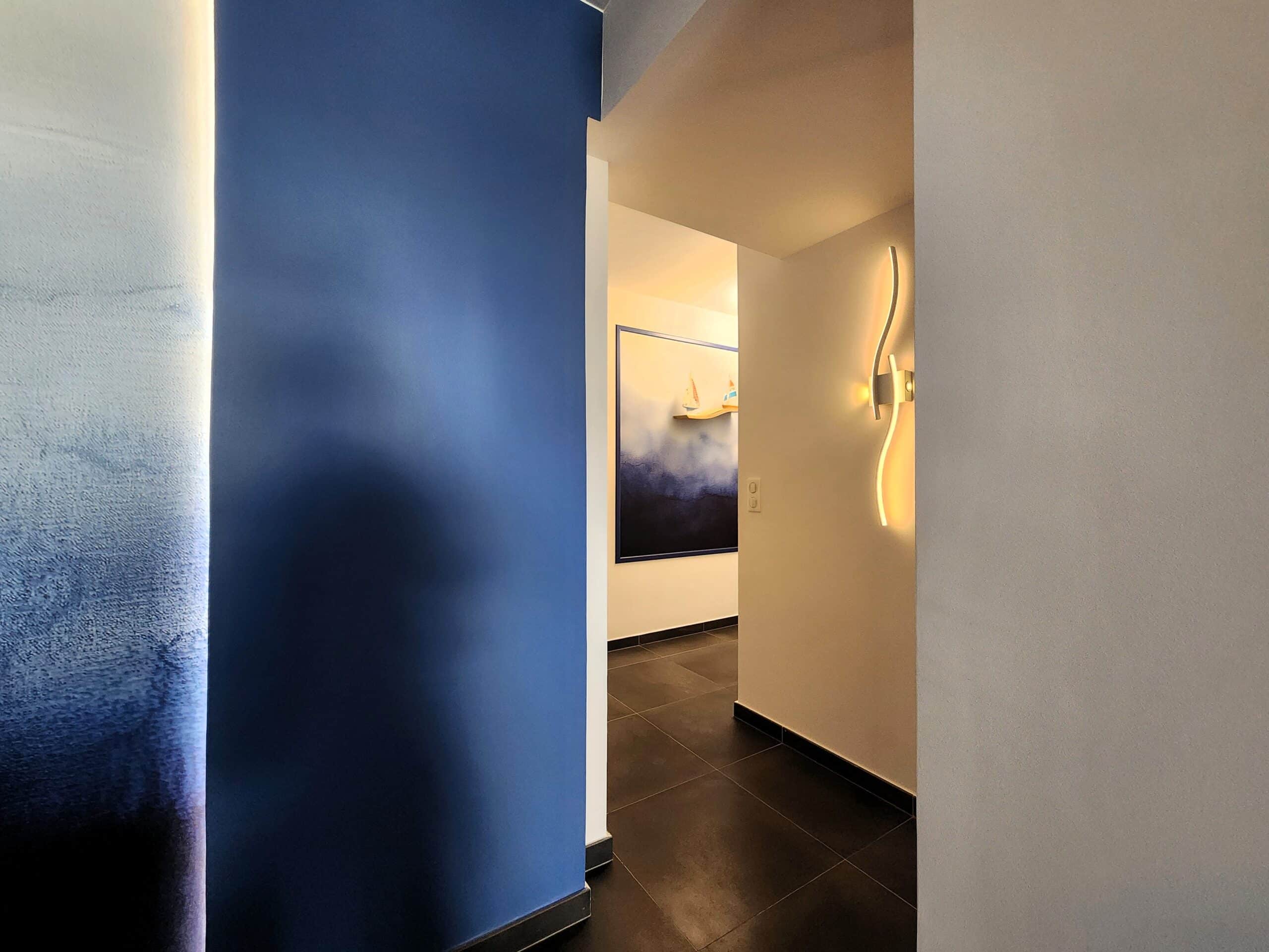 Couloir moderne avec mur bleu, luminaire design ondulé et tableau abstrait dans une décoration intérieure élégante.