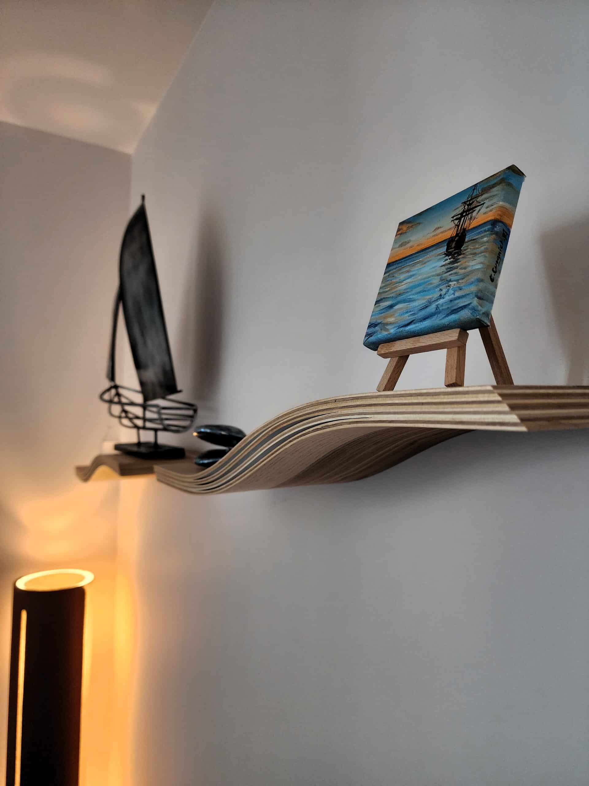 Étagère ondulée en contreplaqué avec un petit tableau sur chevalet et un voilier décoratif sur un mur blanc.