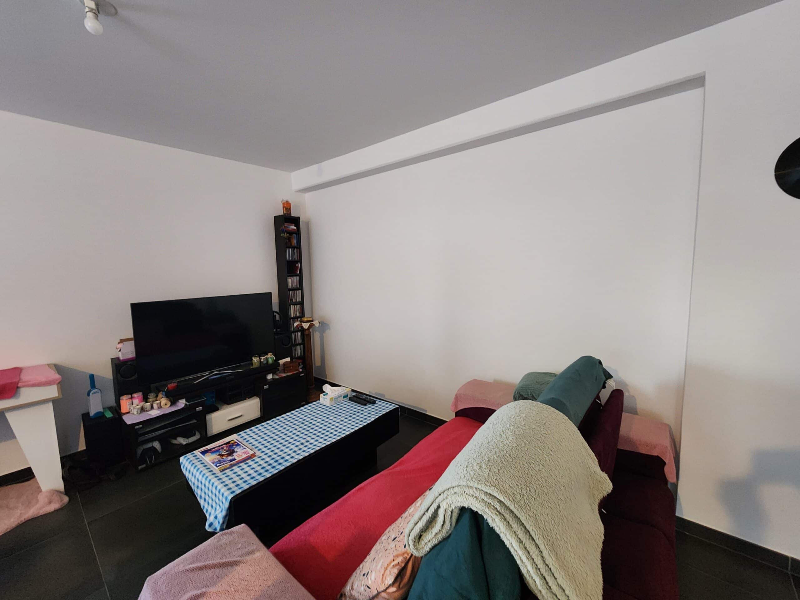 Salon moderne avec canapé rouge, table à carreaux bleus et meuble TV noir devant un grand mur blanc.