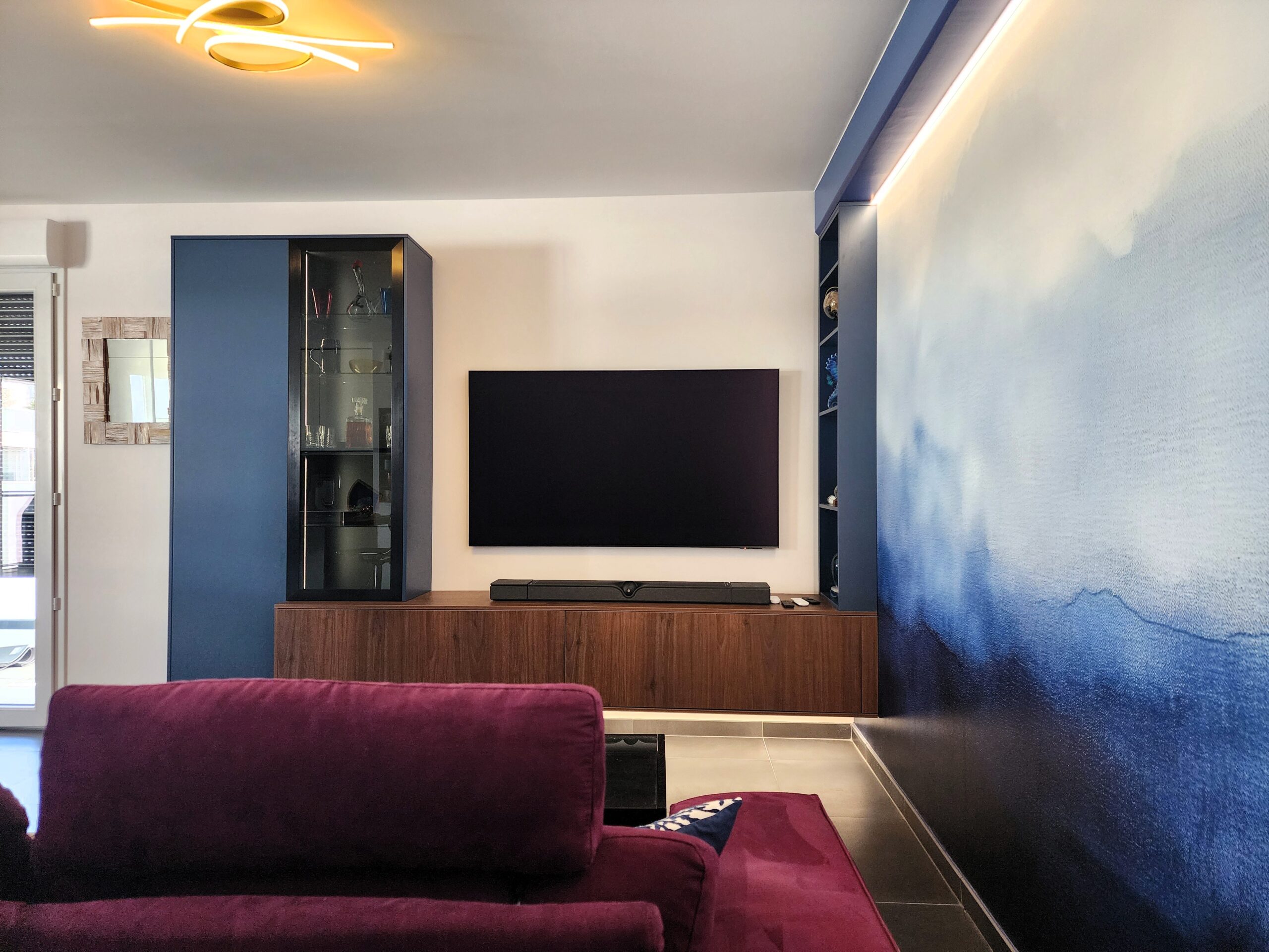 Salon moderne avec canapé violet, meuble TV en bois et mur artistique bleu sous un luminaire design.