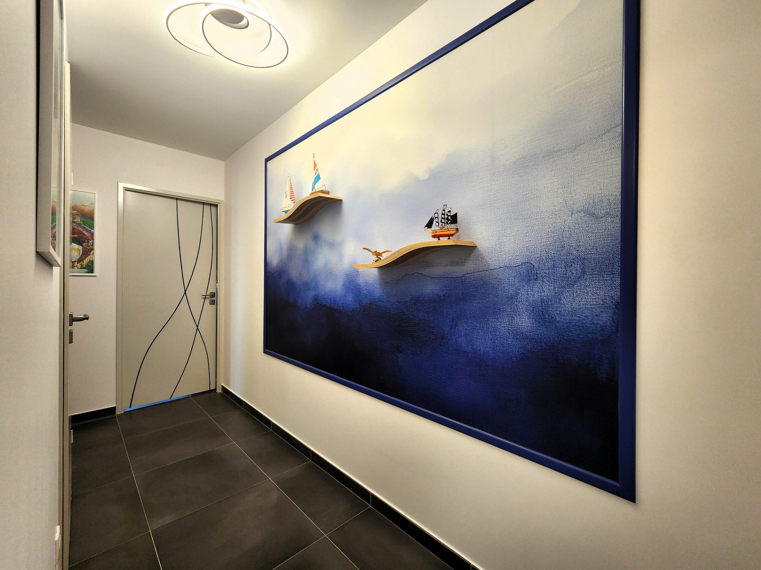 Décoration murale marine avec tableau bleu, étagères vagues et bateaux miniatures dans un couloir moderne.