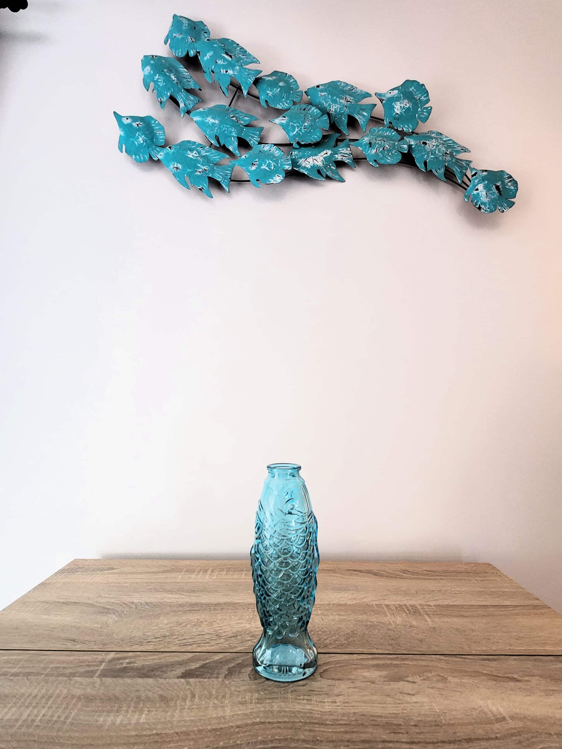 Vase en verre bleu en forme de poisson sur une table en bois sous une sculpture murale de banc de poissons bleus.