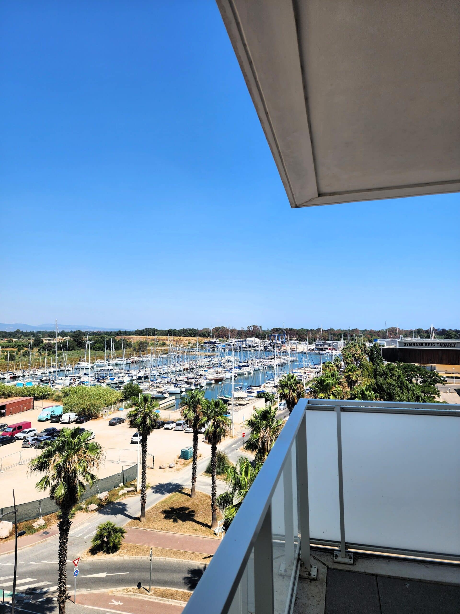Vue d'un balcon sur le port avec palmiers et le Gymnase Éric Tabarly sous un ciel bleu azur ensoleillé.